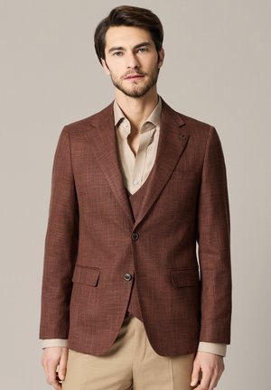 Mężczyzna w brązowej teksturowanej marynarce i pasującej kamizelce, pod którymi ma beżową koszulę wizytową i beżowe spodnie, stojący na jednolitym tle.