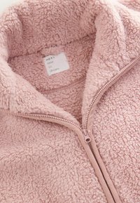 Roze fleecejack met een rits aan de voorkant en een hoge kraag. De stof heeft een zachte, gestructureerde afwerking en toont een verzorgingslabel aan de binnenkant.