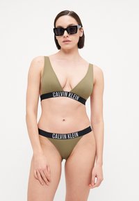 Bikini set u maslinastoj boji s topom na v-izrez i odgovarajućim donjim dijelom niskog struka. Sadrži crni pojas s natpisom "CALVIN KLEIN" u bijelom.