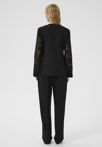 Blazer nero con maniche in pizzo, taglio strutturato e texture liscia. Abbinato a pantaloni neri su misura per un look coeso.