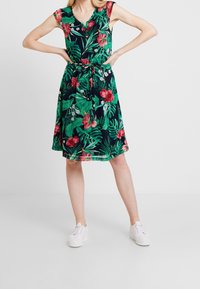 Nainen, joka käyttää olkaimettomaa polvenmittaista mekkoa, jossa on vihreitä lehtiä ja vaaleanpunaisia kukkia, vyötärölle solmittuna, yhdistettynä valkoisiin laskettelutossuihin.