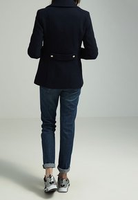 Marineblå uldblandingsjakke med en struktureret silhuet, der har en spidsgår og to metalknapper bagpå, båret over blå jeans og sneakers.