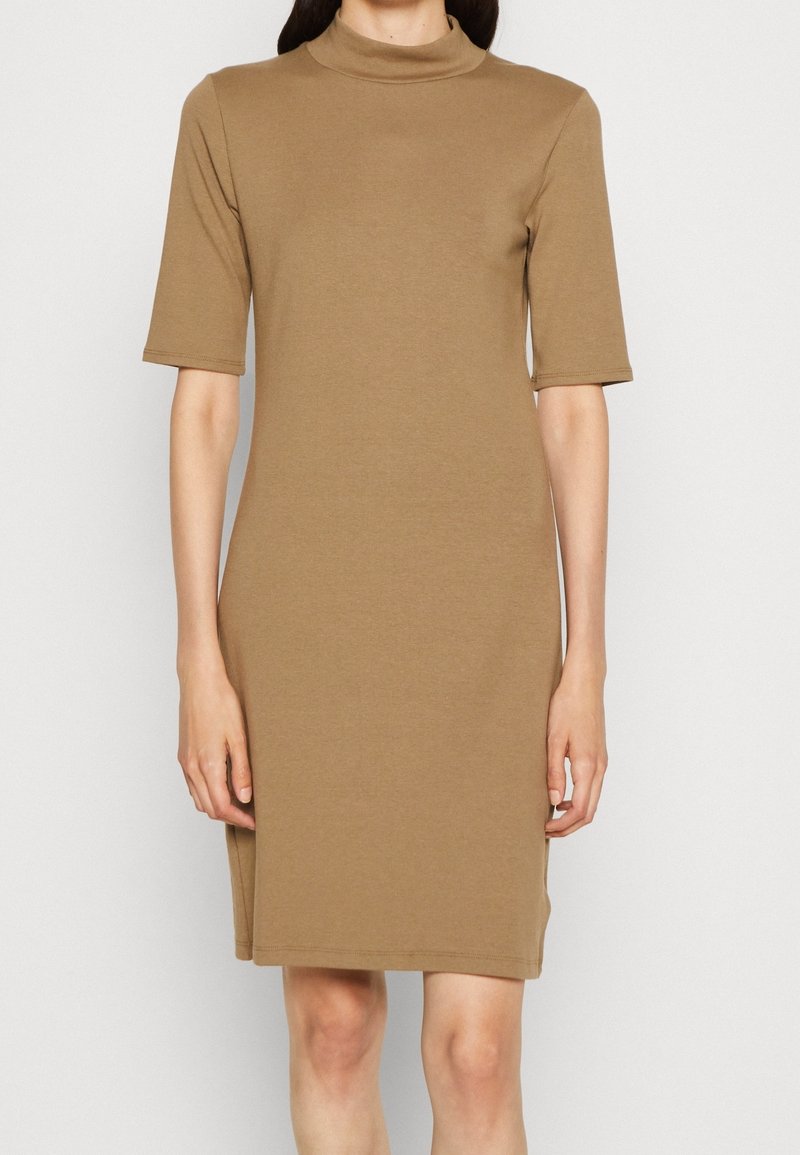 Femme portant une robe marron camel longueur genoux avec des manches aux coudes et un col haut sur un fond clair uni.