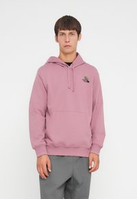 Lyserød hoodie lavet af blødt stof, med en kængurulomme, snøre i hætten og et sort logo på venstre bryst. Stylet med grå bukser.