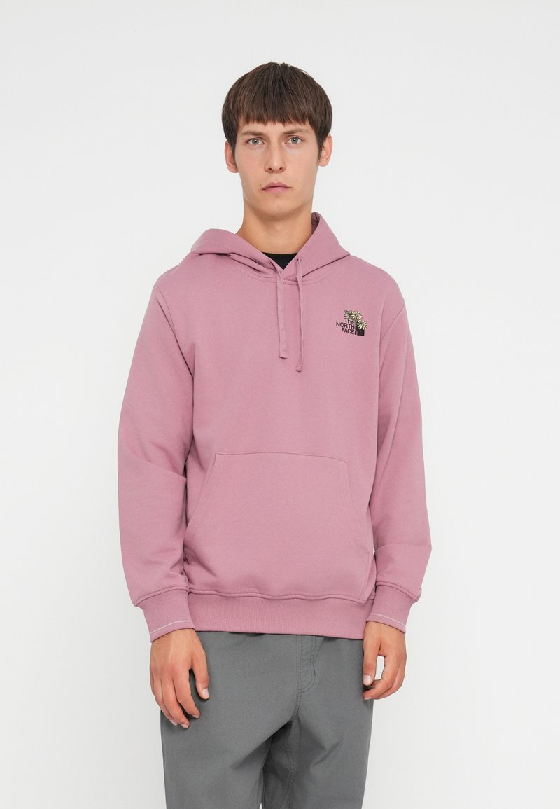 Lyserød hoodie lavet af blødt stof, med en kængurulomme, snøre i hætten og et sort logo på venstre bryst. Stylet med grå bukser.