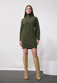 Robe pull en vert olive avec un tissu côtelé et un col haut ; associée à des bottes en suède beige jusqu'au genou.