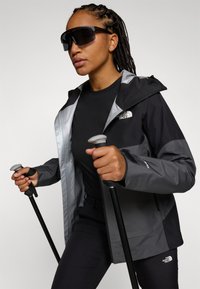 Juodas ir pilkas nepralaidus vandeniui švarkas su užtrauktuku, kapišonu ir "North Face" logotipu; derinamas su juoda marškinėliais ir žygio lazdomis; elegantiškas, modernus dizainas.