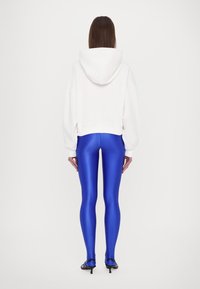 Hvit cropped hettegenser med hette, kombinert med skinnende, blå leggings. Leggingsene er tettsittende og glansfulle, og antrekket inkluderer svarte hælede sandaler.
