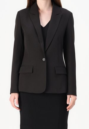 Femme portant un blazer noir sur mesure avec un bouton décoratif unique et des poches à rabat, sur un haut et une jupe noirs.