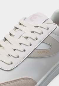 Witte leren sneaker met witte veters, beige suède neuskapje en de tekst "COURT '79" op het zijpaneel.