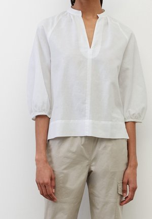 Personne portant un chemisier blanc à manches longues avec un col en V et un pantalon beige avec une poche latérale, debout devant un fond uni.