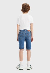 Modré denimové kraťasy s rovnými nohavicemi, s zadními kapsami a hnědou nášivkou na pásku, nošené s bílým tričkem a botami.