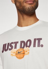 Hvit bomulls t-skjorte med teksten "JUST DO IT.", en oransje basketballgrafikk og blomsterdetaljer i myke toner.