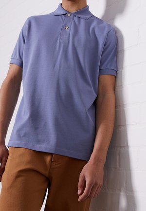 Man wearing a short-sleeve light purple polo shirt and brown pants, standing against a white brick wall.