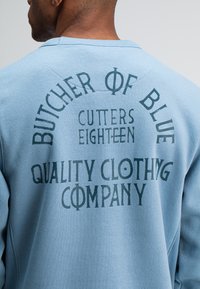 Lichtblauwe sweatshirt van een textuurstof, met opvallende groene typografie op de achterkant. De tekst bevat "BUTCHER OF BLUE" en "QUALITY CLOTHING COMPANY."