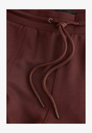 Pantalon de survêtement bordeaux avec une taille à cordon de serrage, présentant une texture lisse et des embouts marron contrastants sur les cordons.