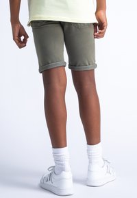 Grønne denimshorts med optrukne bukseben, parret med hvide sneakers og tykke hvide sokker, set fra ryggen. Glat tekstur, afslappet stil.