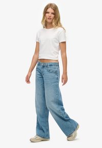 Biały krótki t-shirt w połączeniu z jasnoniebieskimi spodniami typu wide-leg, z zapięciem na guziki i subtelnymi przetarciami. Na nogach beżowe sneakersy.