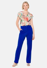 Bloemmotief korte mouw blouse met een V-hals, gecombineerd met hoogsluitende, rechte koningsblauwe broeken. Model draagt zilveren sandalen.