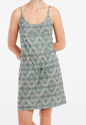 Vrouw draagt een mouwloze jurk tot op de knie met groene en witte geometrische bloemmotieven en dunne schouderbandjes.