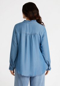 Camisa de mezclilla azul claro con mangas largas, cuello con volantes y detalle de espalda fruncida. Cuenta con puños abotonados y un dobladillo curvado.