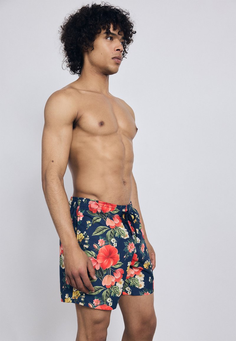 Jeune homme aux cheveux frisés portant un short de bain à imprimé floral bleu marine, se tenant torse nu devant un fond gris clair uni.