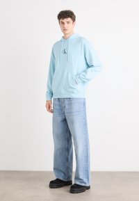 Sudadera de algodón azul claro con un bolsillo frontal, capucha con cordones y detalle de logo. Combinada con vaqueros anchos desgastados de color azul y zapatos negros de plataforma.