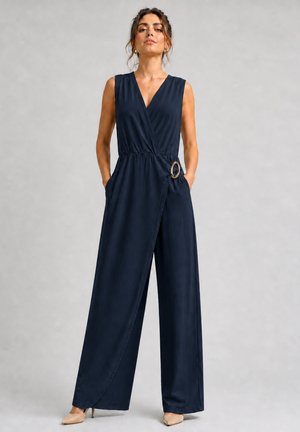 Femme portant une combinaison cache-cœur sans manches bleu marine avec jambes larges, une boucle à la taille, les mains dans les poches et des talons hauts beige, debout avec assurance.