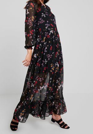 Femme portant une longue robe noire transparente avec un motif floral coloré et des sandales noires à talons blocs ouvertes sur un fond uni.