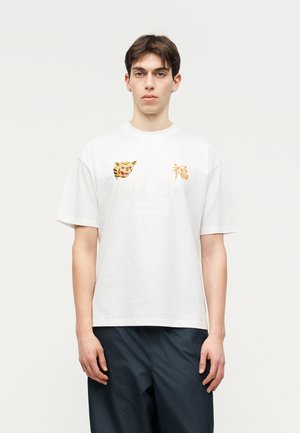 Ung mand stående, iført hvid T-shirt med tigermotiv og broderede asiatiske tegn, kombineret med mørke, løse bukser.