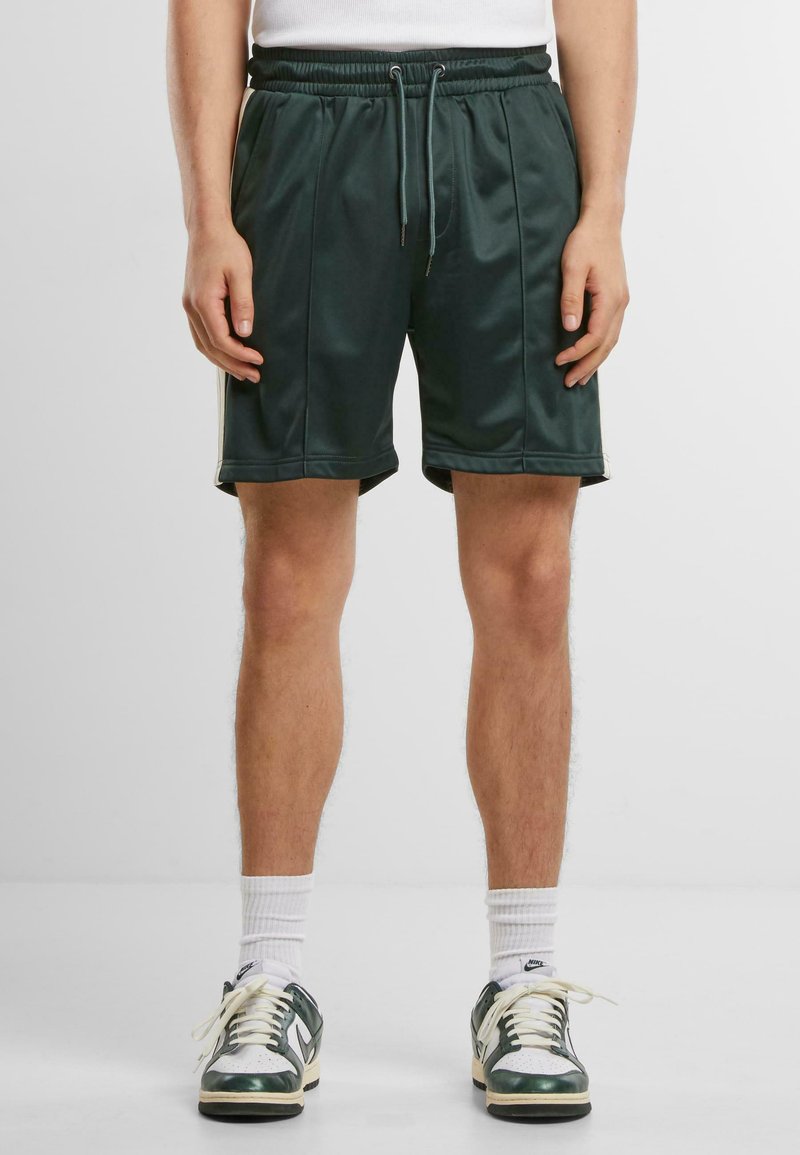 Shorts de sport vert foncé avec une taille élastique, un cordon de serrage et une texture lisse. Associés à des baskets blanches et des chaussettes mi-mollet.
