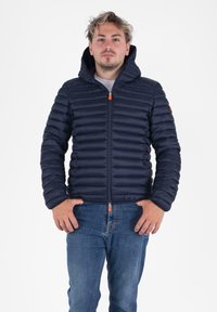 Jonge man in een marineblauwe gewatteerde hoodie en blauwe spijkerbroek, staande met zijn handen bij zijn zakken tegen een witte achtergrond.