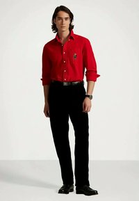 Fiatal férfi áll, piros gombos ingben emblémával, fekete nadrágban, fekete övben, fekete cipőben és karórában.