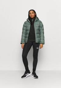 Grøn pufferjakke med hætte, syntetisk materiale, ribbede manchetter; sort tætsiddende sweater, sorte leggings med logo og sorte atletiske sko.