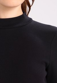 Musta ribbineulostettu paita, jossa korkea kaulus ja teksturoitu kangas, pystysuuntaisilla viivoilla ja vartalonmyötäisellä siluetilla.