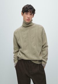 Beigefarbener Rollkragenpullover mit Zopfmuster und geripptem Kragen, kombiniert mit dunkelbraunen Cordhosen. Glatte Textur und lockere Passform.