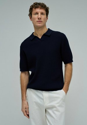 Hombre vistiendo una camiseta polo de punto de manga corta azul oscuro y pantalones blancos, de pie con una mano en el bolsillo contra un fondo liso.