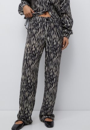 Femme portant un pantalon à imprimé zèbre noir et beige et un haut à manches longues assorti, avec des ballerines noires à bout carré sur un fond uni.