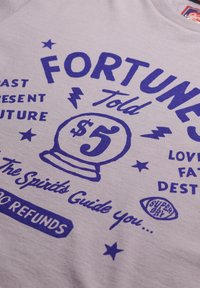 Camiseta gris claro con texto gráfico audaz en azul e ilustraciones relacionadas con la adivinación, incluyendo una bola de cristal y un signo de dólar.