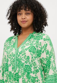 Πράσινο φόρεμα με λουλουδάτο μοτίβο, v-προσοχή, μακριά μανίκια και tiered σχέδιο. Διαθέτει λεπτομερή κεντήματα κατά μήκος της λαιμόκοψης και των μανικιών.