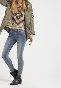 Olivgrön militärjacka över en beige mönstrad tröja, kombinerad med blå smala jeans och svarta ankelskor med gulddetaljer.