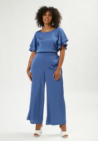 Blauwe, zijdeachtige jumpsuit met gerimpelde mouwen, wijd uitlopende broekspijpen en een getailleerde taille. Gecombineerd met witte hakken, tegen een neutrale achtergrond.