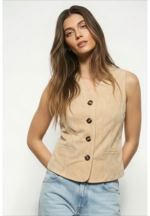 Kvinde med langt brunt hår iført en beige ærmeløs vest med knapper og lyseblå jeans, poserer mod en ensfarvet baggrund.