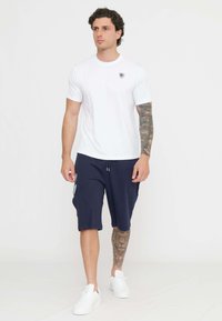 T-shirt bianca in cotone con maniche corte, pantaloni corti blu navy con tasche laterali e dettaglio del logo, e sneakers bianche. Texture liscia, design casual.