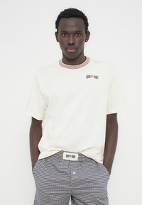 Κρεμ μπλούζα με κόκκινο ριμπ γιακά, με κεντημένο το λογότυπο "HOUSE OF SUNNY" σε κόκκινο. Συνδυάζεται με ναυτικό μπλε και λευκό καρό σορτς.