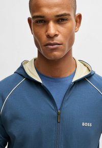 Blue zip-up hoodie with cream accents, made from soft fabric. Features a front zip, attached hood, and 'BOSS' logo on the chest.