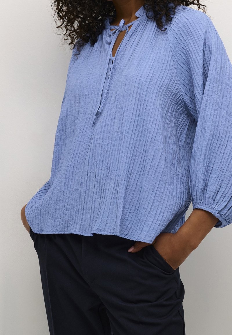 Blouse en coton bleu clair texturé avec une fermeture à nouer, une patte boutonnée et des manches ballon, associée à un pantalon foncé.