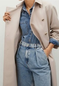 Beige trenchcoat met knopen, gedragen over een blauwe denim shirt en lichtblauwe high-waisted jeans, met voorzakken en een relaxte pasvorm.