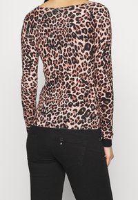 Femme portant un haut à manches longues imprimé léopard rose et noir, associé à un jean noir ajusté, vue de dos sur un fond neutre.