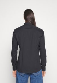 Zwarte lange mouwen shirt met een puntige kraag, knopenmanchetten en een gladde textuur, gedragen met een blauwe spijkerbroek, van achteren bekeken.